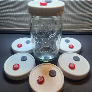 May include: A clear glass jar with a white lid and several white jar lids with red and gray components. The jar is surrounded by the lids, suggesting a set for a specific purpose. The background features a metallic, slatted structure.
