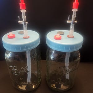 May include: Two clear glass Ball jars with light blue lids. Each jar has a clear tube with a red cap and a small red cap on the lid. The jars are on a brown surface against a dark background.
