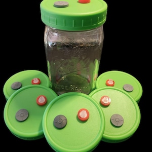May include: A clear glass jar with a green lid, surrounded by several green lids. Each lid has a gray and a red component. The jar and lids are likely for food preservation or storage.