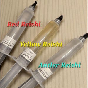May include: Three clear syringes filled with different colored liquids, labeled "Red Reishi", "Yellow Reishi", and "Antler Reishi". The syringes are on a light brown wooden surface. The text is in a colorful font.