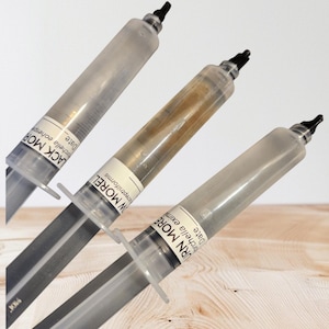May include: Three clear syringes with black tips, each containing a liquid. The syringes have white labels with black text that reads "BURN MORE" and "Morchella eximia". The liquid in one syringe is amber-colored.