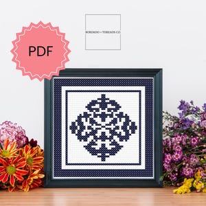 May include: A printable PDF cross stitch pattern featuring a geometric design in black and white. The pattern is framed in a dark blue frame and displayed on a wooden table.
