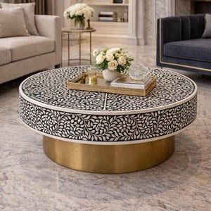 Puede incluir: Una mesa de centro redonda con un patrón floral incrustado en blanco y negro en la parte superior y los laterales, apoyada sobre una base cilíndrica de metal dorado. Una bandeja rectangular dorada sobre la mesa sostiene un jarrón blanco con flores.