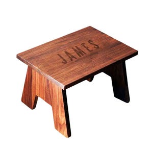 Puede incluir: Un pequeño taburete de madera con una parte superior rectangular y patas anguladas. El taburete es de madera marrón oscuro y presenta el nombre "JAMES" grabado en la superficie superior. El diseño es simple y funcional.