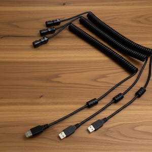 Może przedstawiać: Zestaw trzech czarnych kabli spiralnych, z których każdy posiada złącze USB-A na jednym końcu i złącze cylindryczne na drugim. Każdy kabel zawiera mały, czarny cylindryczny element ferrytowy umieszczony na prostym odcinku przewodu.
