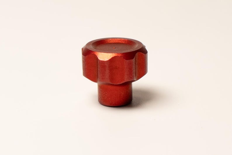 Puede incluir: Un pomo de metal rojo con un borde acanalado y una base cil&iacute;ndrica. El pomo tiene una superficie texturizada para el agarre y probablemente se utiliza para ajustar o apretar. El objeto est&aacute; sobre un fondo blanco liso.