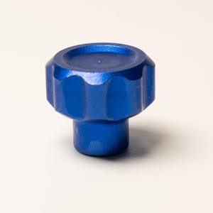 Puede incluir: Un pomo azul brillante con una parte superior circular estriada y una base cil&iacute;ndrica. El pomo est&aacute; hecho de un material s&oacute;lido y est&aacute; sobre un fondo blanco. El dise&ntilde;o sugiere un elemento de hardware funcional o decorativo.