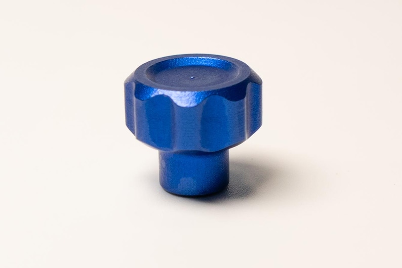 Puede incluir: Un pomo azul brillante con una parte superior circular estriada y una base cil&iacute;ndrica. El pomo est&aacute; hecho de un material s&oacute;lido y est&aacute; sobre un fondo blanco. El dise&ntilde;o sugiere un elemento de hardware funcional o decorativo.