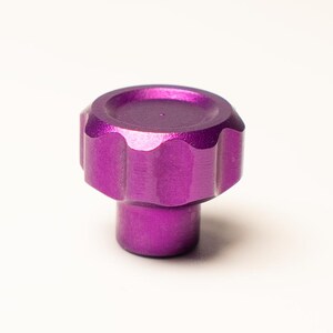 Puede incluir: Primer plano de un pomo morado con borde acanalado y base cil&iacute;ndrica. El pomo es de metal y tiene un acabado mate. El fondo es blanco liso.