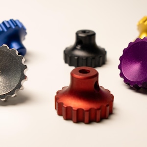 Peut inclure: Un assortiment de boutons métalliques usinés de couleurs vives : argent, bleu, noir, rouge, violet et or. Chaque bouton a un dessus circulaire et strié et une base cylindrique avec un trou. Les boutons sont disposés sur une surface blanche.
