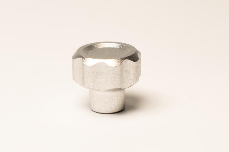 Puede incluir: Primer plano de un pomo plateado con borde estriado y base cil&iacute;ndrica. El pomo tiene una parte superior ligeramente c&oacute;ncava y est&aacute; sobre un fondo blanco. El objeto parece ser de metal.