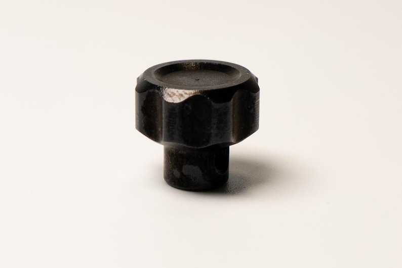 Puede incluir: Un pomo oscuro y texturizado con una parte superior redondeada y un borde acanalado en forma de engranaje. El pomo est&aacute; hecho de un material oscuro, posiblemente met&aacute;lico, y se asienta sobre una superficie blanca. El dise&ntilde;o sugiere una est&eacute;tica industrial funcional.