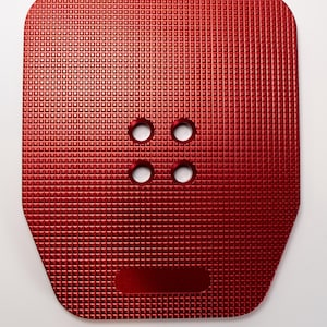 Puede incluir: Placa metálica roja con un patrón texturizado en forma de rejilla. Cuatro agujeros circulares están cerca del centro y una hendidura rectangular en la parte inferior. La placa tiene esquinas redondeadas.