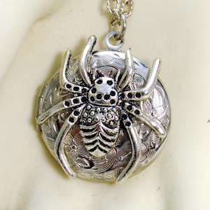 May include: Silver-toned spider pendant necklace. The pendant features a detailed spider design atop a round, engraved locket. The spider has a textured body and legs. The necklace chain is also silver.