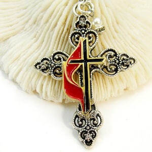 May include: Silver-toned cross pendant necklace with a red and black flame design. The cross features ornate filigree detailing and small clear crystals. A small pearl charm hangs near the top of the cross. The necklace is displayed on a white, textured surface.