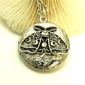 May include: A round silver-toned metal locket pendant suspended from a matching chain. The locket features a raised relief design of a moth with crescent moon patterns on its wings, set against a background of engraved floral and swirling leaf motifs.