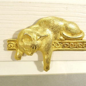 May include: A gold-toned metal decorative accent featuring a cat lying down on a horizontal bar with a scrollwork pattern. The cat is depicted in a resting position with one front paw hanging over the edge of the bar and a textured finish.