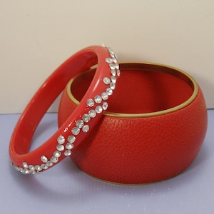 May include: A thin, glossy red bangle bracelet adorned with a single row of clear, sparkling rhinestones, positioned next to a wider, textured red bangle bracelet featuring a smooth, metallic gold-toned interior rim and a pebbled exterior surface.