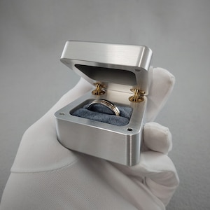May include: A silver ring box with a hinged lid, open to reveal a silver and gold ring resting on a gray velvet cushion.