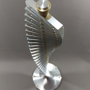 May include: A silver and gold metal sculpture with a spiral design. The sculpture is made of many small, flat pieces of metal that are stacked on top of each other. The top of the sculpture has a round, silver knob.