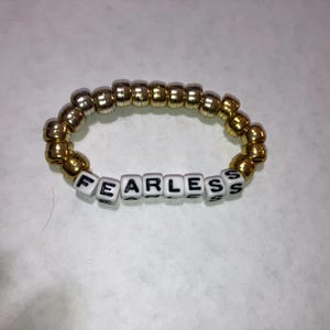 May include: A gold and silver beaded bracelet with the word "FEARLESS" spelled out in white letter beads.