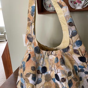 May include: A large, yellow canvas tote bag with a floral pattern in shades of blue, brown, and beige. The bag has a long strap and a wide opening.