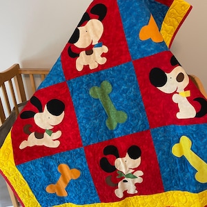 May include: A quilted blanket featuring a repeating pattern of red and blue squares. The red squares display cartoon dogs with black ears and colorful collars, while the blue squares feature yellow and green dog bones. A yellow border frames the quilt.