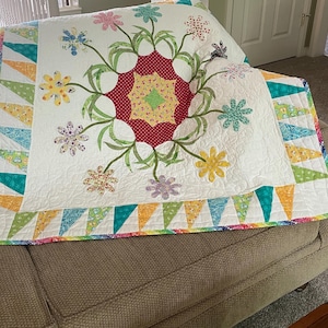 May include: A handmade quilt with a floral design. The quilt features a white background with a central red and yellow floral pattern. Colorful flowers and green leaves are scattered across the quilt. The border is made of colorful triangles.
