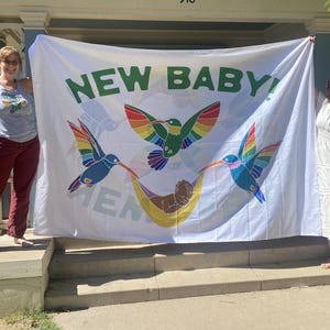 May include: White banner with the text "NEW BABY!" in green letters. The banner features three colorful hummingbirds with rainbow wings and a baby in a hammock.