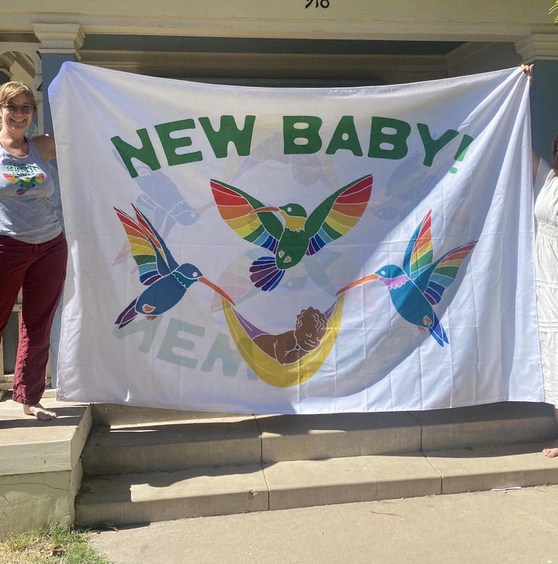 May include: White banner with the text "NEW BABY!" in green letters. The banner features three colorful hummingbirds with rainbow wings and a baby in a hammock.