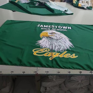 May include: Green banner with the text "Jamestown High School" and a white eagle with yellow accents. The text "Eagles" is written in a yellow script font below the eagle.