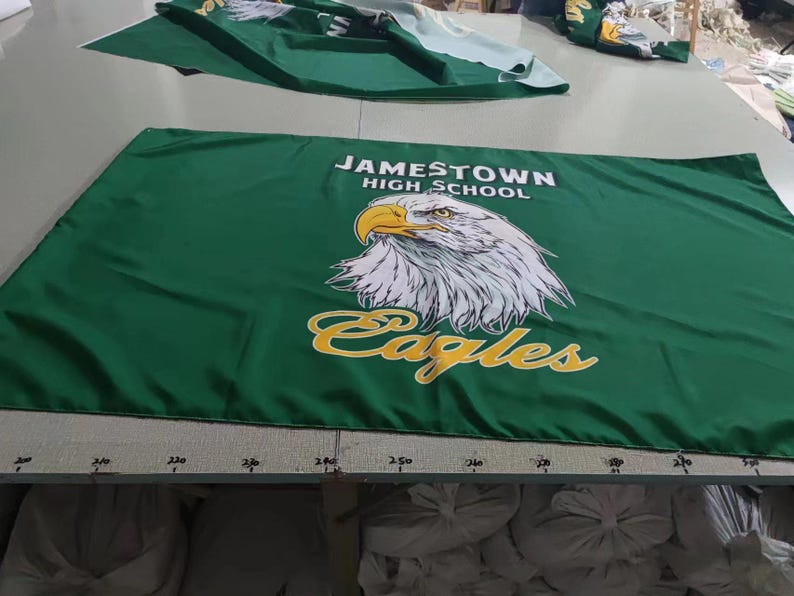 May include: Green banner with the text "Jamestown High School" and a white eagle with yellow accents. The text "Eagles" is written in a yellow script font below the eagle.