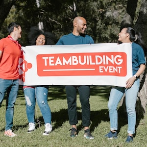 May include: Four people holding a white banner with red text that says "Team Building Event". The people are smiling and looking at each other. They are standing in a grassy area with trees in the background.