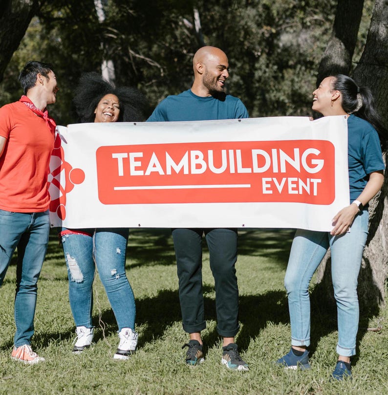 May include: Four people holding a white banner with red text that says "Team Building Event". The people are smiling and looking at each other. They are standing in a grassy area with trees in the background.