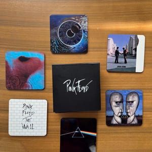 Puede incluir: Un conjunto de posavasos temáticos de Pink Floyd con ilustraciones de álbumes. Los posavasos muestran imágenes de un ojo, dos hombres dándose la mano, dos cabezas de piedra, un prisma y la portada del álbum "The Wall". También hay una caja negra con el logotipo de la banda.