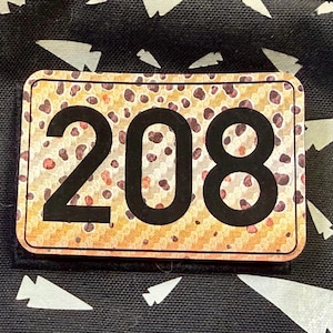 May include: A rectangular patch featuring the black number 208 centered on a tan and brown leopard print background with a thin black border. The patch is attached to a black fabric surface decorated with a repeating pattern of white arrowheads.