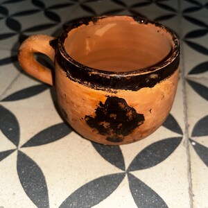 May include: A small, round, terracotta-colored clay mug featuring a thick, dark brown painted rim and a matching dark brown abstract, cross-like shape painted on the front. The mug has a single, curved handle attached to the side.