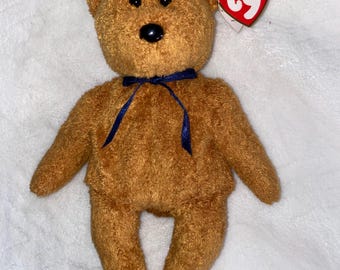 May include: A brown Beanie Baby teddy bear with a blue ribbon around its neck. The bear has a red and yellow tag that says "Beanie Babies" and "ty".