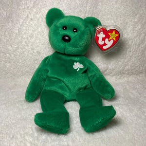 May include: Green plush beanie baby bear with a black nose and a white shamrock on its chest. The bear is sitting on a white fuzzy surface. The bear has a Ty tag attached to its right ear.