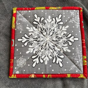 Peut inclure: Un dessous de plat carré et matelassé de Noël avec un grand motif de flocon de neige blanc sur fond gris. Le dessous de plat a une bordure rouge avec une garniture verte et jaune. Le flocon de neige est détaillé avec des coutures blanches.