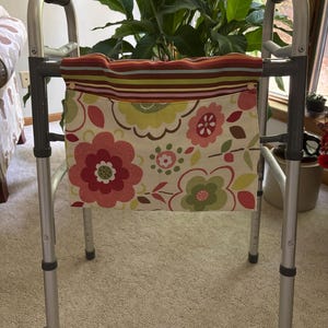 May include: A silver walker with a colorful floral patterned bag attached. The bag features large red and pink flowers, green leaves, and a striped top border. The walker has black hand grips and wheels.