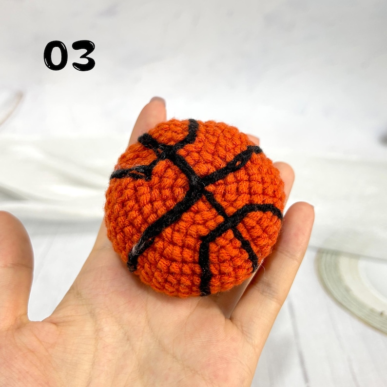 May include: A small, round, orange crocheted basketball with black yarn detailing representing the seams. The number 03 is printed in black in the upper left corner of the image, indicating the item number for this specific sports-themed toy.