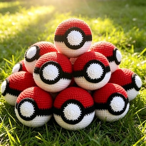 May include: A pile of red, white, and black crocheted balls, resembling Pokeballs, are stacked on green grass. The balls have a white center with a black outline and a black stripe. The scene is lit by sunlight.