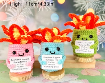 May include: Three crocheted plush plants in pots, each with a sign that reads "Emotional Support Dumpster Fire It's fine, I'm fine. Everything is fine!" The plants are orange, blue, and green. The pots are pink, blue, and green. The plants are 11 cm tall.