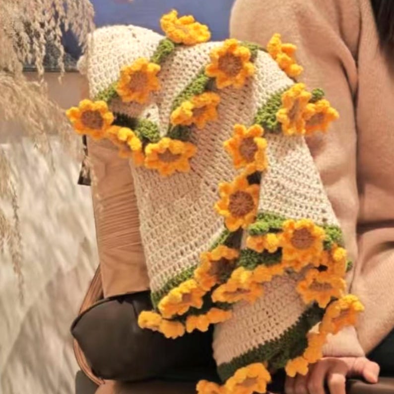 K&ouml;nnte beinhalten: Eine geh&auml;kelte Decke mit einem Sonnenblumenmuster. Die Decke ist aus beige und gr&uuml;nem Garn gefertigt und hat einen Rand aus geh&auml;kelten Sonnenblumen.