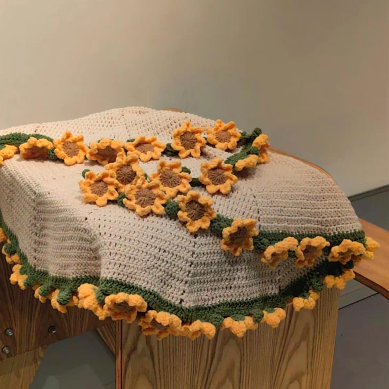 K&ouml;nnte beinhalten: Eine geh&auml;kelte Decke mit einem Sonnenblumenmuster. Die Decke ist beige mit gr&uuml;nen Akzenten und einem Rand aus gelben geh&auml;kelten Sonnenblumen.