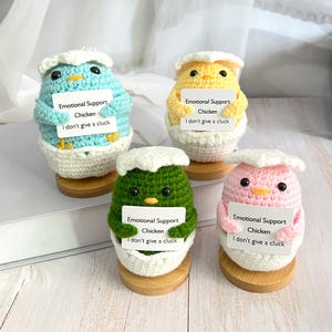 May include: Four crocheted chicks in eggshells, each holding a sign that reads "Emotional Support Chicken I don't give a cluck". The chicks are blue, yellow, green, and pink.