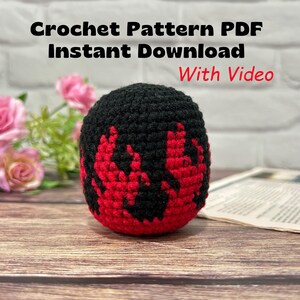 May include: A spherical crochet object featuring a black base with a red flame pattern. The text above reads Crochet Pattern PDF Instant Download With Video. The item is a handmade textile craft piece with a dense, textured yarn construction.