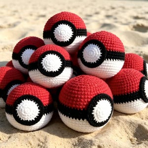 May include: A pile of red, white, and black crocheted balls, resembling Pokeballs, are arranged on a sandy surface. The balls have a red top, a white center with a black ring, and a black stripe. The balls are of varying sizes.