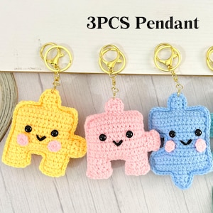 Peut inclure: Trois porte-clés en forme de pièce de puzzle au crochet, jaunes, roses et bleus, chacun avec un visage souriant et attaché à un porte-clés doré. Le texte "3PCS Pendant" est affiché au-dessus des porte-clés.